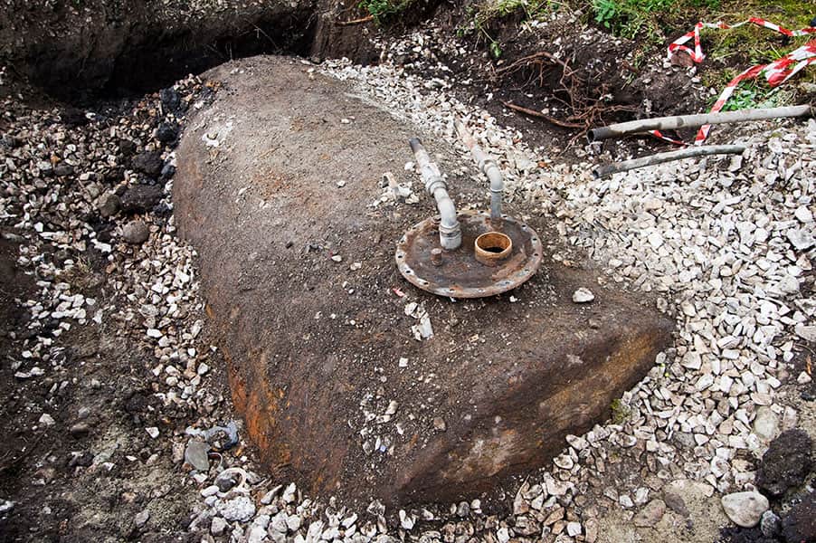 Un serbatoio di metallo arrugginito parzialmente interrato in un cantiere. Le tubazioni sporgono dalla parte superiore, circondato da ghiaia e terreno smosso.