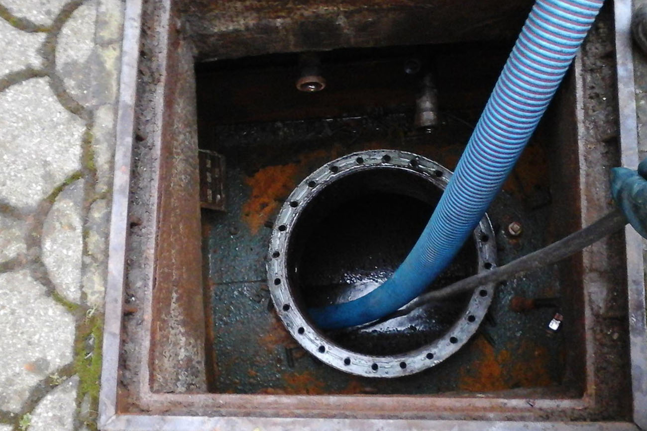 Immagine che mostra un pozzetto di ispezione con tubo flessibile blu in fase di aspirazione. La superficie interna è macchiata di ruggine e acqua. Fase di manutenzione.
